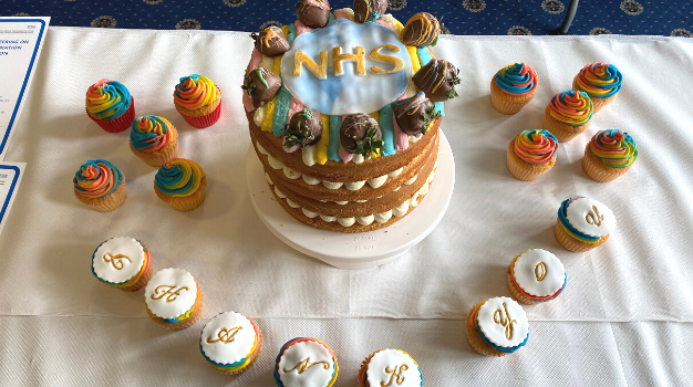 A celebration cake with rainbow decorations and 'NHS' written on the top, along with cupcakes that spell out the words 'thank you'